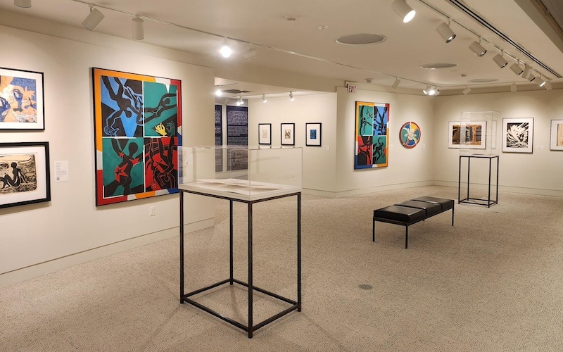 A gallery view with colorful framed artworks on the walls and two display cases exhibiting booklets and a leather bench in the middle of the gallery