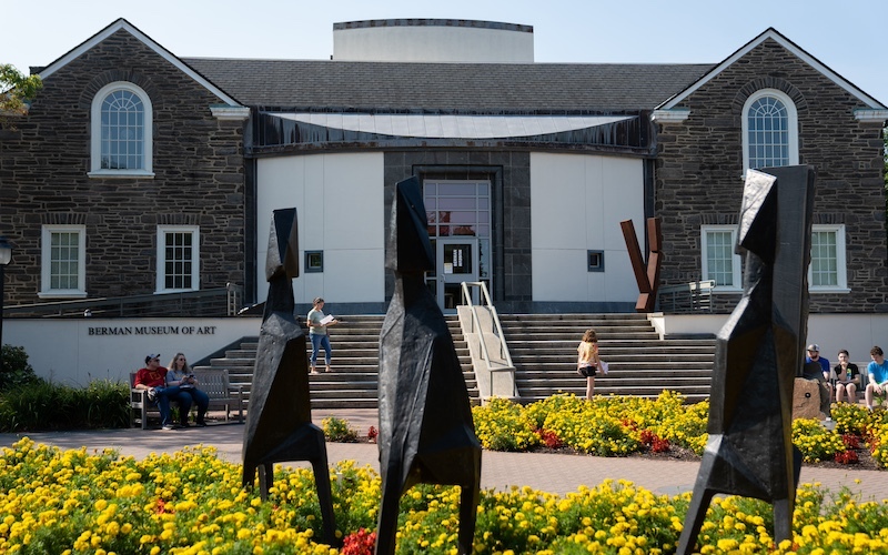 three black figure-looking sculptures stand in a bed of yellow flowers with a stone building with steps leading to entrance in background. people are walking on steps and sitting on benches flanking the stairs