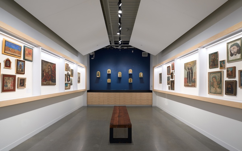 white room with framed old depictions of saints on left and right wall; small works of art on a dark blue wall at the back of the room; a bench is in the middle of the room