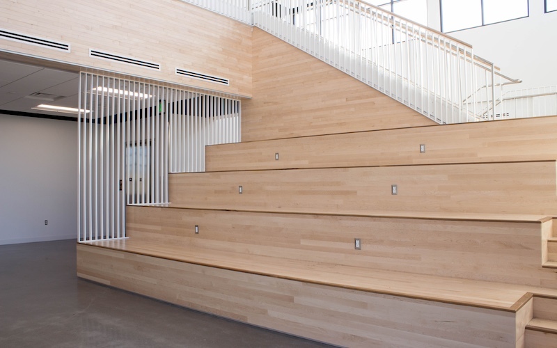 light wooden bleachers in the middle of large white room with windows on second level and a staircase in the background