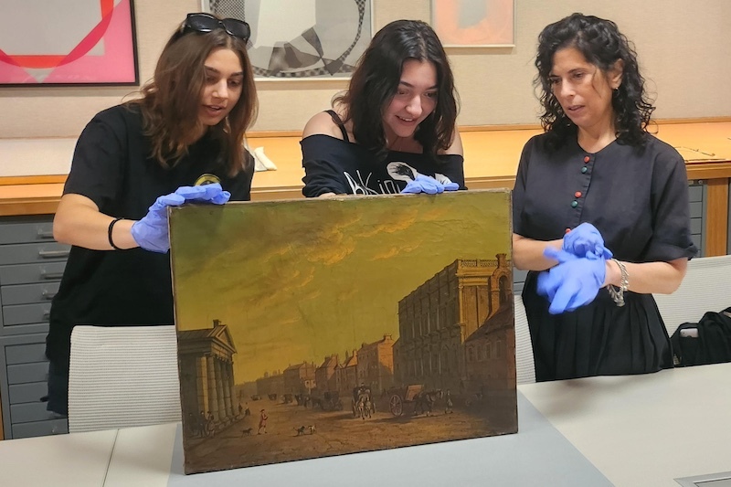 two female students stand behind an unframed painting of an old town landscape, propping it up with gloved hands. Another woman stands to the side watching